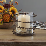 Barbed Wire Pillar Candle Rustic Holder featuring a clear glass cylinder wrapped with dark brown barbed wire, containing a lit cream pillar candle and small decorative stones, set on a wooden tabletop with fall florals