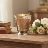 Fleur de Lys Tumbler Hand-Poured Soy Scented Candle - Champagne lit candle in an ornate, textured gold and silver-toned glass holder with a footed base, shown on a table with a stack of books and white roses with baby's breath.