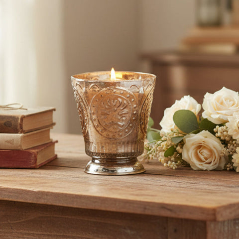 Fleur de Lys Tumbler Hand-Poured Soy Scented Candle - Champagne lit candle in an ornate, textured gold and silver-toned glass holder with a footed base, shown on a table with a stack of books and white roses with baby's breath.