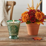 Fleur de Lys Green Tumbler Hand-Poured Soy Scented Candle is a lit green embossed glass candle and a terracotta pot of autumn flowers with orange, red, and yellow blooms sit on a wooden table in a softly blurred indoor setting.
