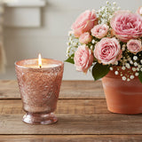 Fleur de Lys Pink Tumbler Hand-Poured Soy Scented Candle in a textured, rose pink-silver-toned glass holder sits on a rustic wooden table next to a terracotta pot of pink roses and baby's breath.