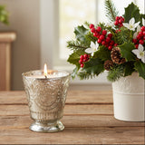 Fleur de Lys Silver Tumbler Hand-Poured Soy Scented Candle with embossed floral detailing sits on a rustic wooden table beside a white vase filled with holly leaves, red berries, pinecones, and white flowers.