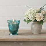 Fleur de Lys Turquoise Tumbler Hand-Poured Soy Scented Candle is a teal blue textured glass candle holder with a lit candle sits on a rustic wooden shelf next to a ceramic vase filled with white and cream roses and green leaves, set against a white paneled wall.