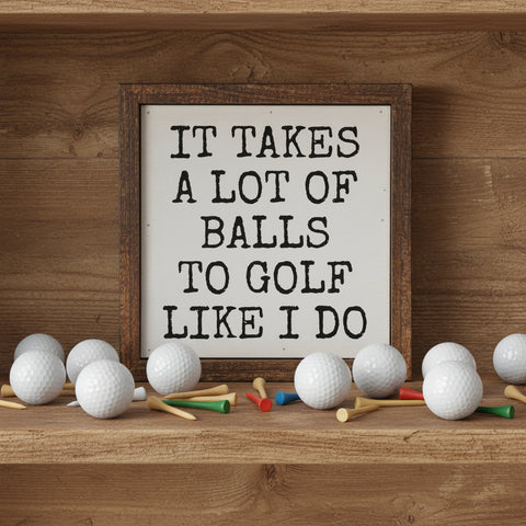 A square framed decorative sign with a rustic brown wood frame and a white face on a wood shelf with golf balls and golf tees. The sign features the humorous text 'IT TAKES A LOT OF BALLS TO GOLF LIKE I DO'.