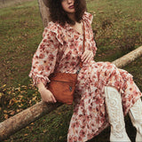 Woman in a floral dress and white boots sitting on a log in a field with trees in the background