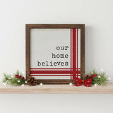 Our Home Believes Vintage Christmas Sign 6x6 A square framed wall sign with a rustic dark brown wooden frame. The sign features a white background with the typewriter-style text 'our home believes' in black, accented by red vertical and horizontal stripes on the right and bottom edges. Displayed on a wood shelf with pine boughts and red ribbon.