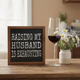 A square wooden sign with a dark chalkboard-style center and distressed white text that reads, 'RAISING MY HUSBAND IS EXHAUSTING.'.