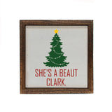 A square framed decorative sign with a rustic dark brown wood frame. The sign has an off-white background featuring a green Christmas tree with a yellow star and the red text 'SHE'S A BEAUT CLARK.