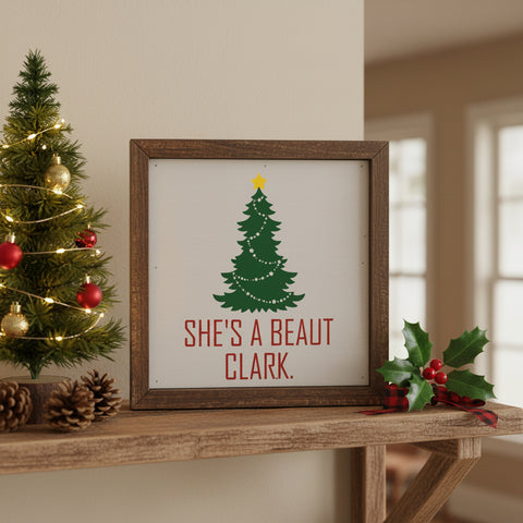 A square framed decorative sign with a rustic dark brown wood frame. The sign has an off-white background featuring a green Christmas tree with a yellow star and the red text 'SHE'S A BEAUT CLARK. Displayed next to a mini Christmas tree with pinecones and holly on a shelf.