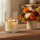 Artisan Clear Bubble Glass Hurricane Hand-Poured Soy Scented Candle in a textured clear glass holder sits on a wooden table beside a ceramic vase filled with autumn flowers, in a softly blurred indoor setting.